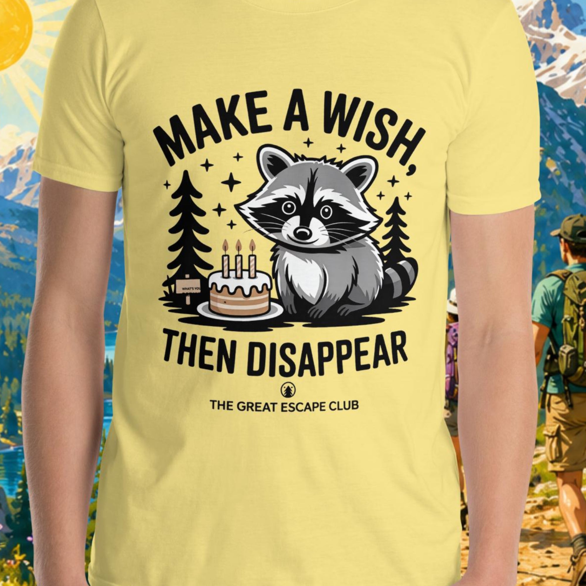 Person wearing a yellow t-shirt with a raccoon graphic and text in front of a scenic mountain backdrop.
