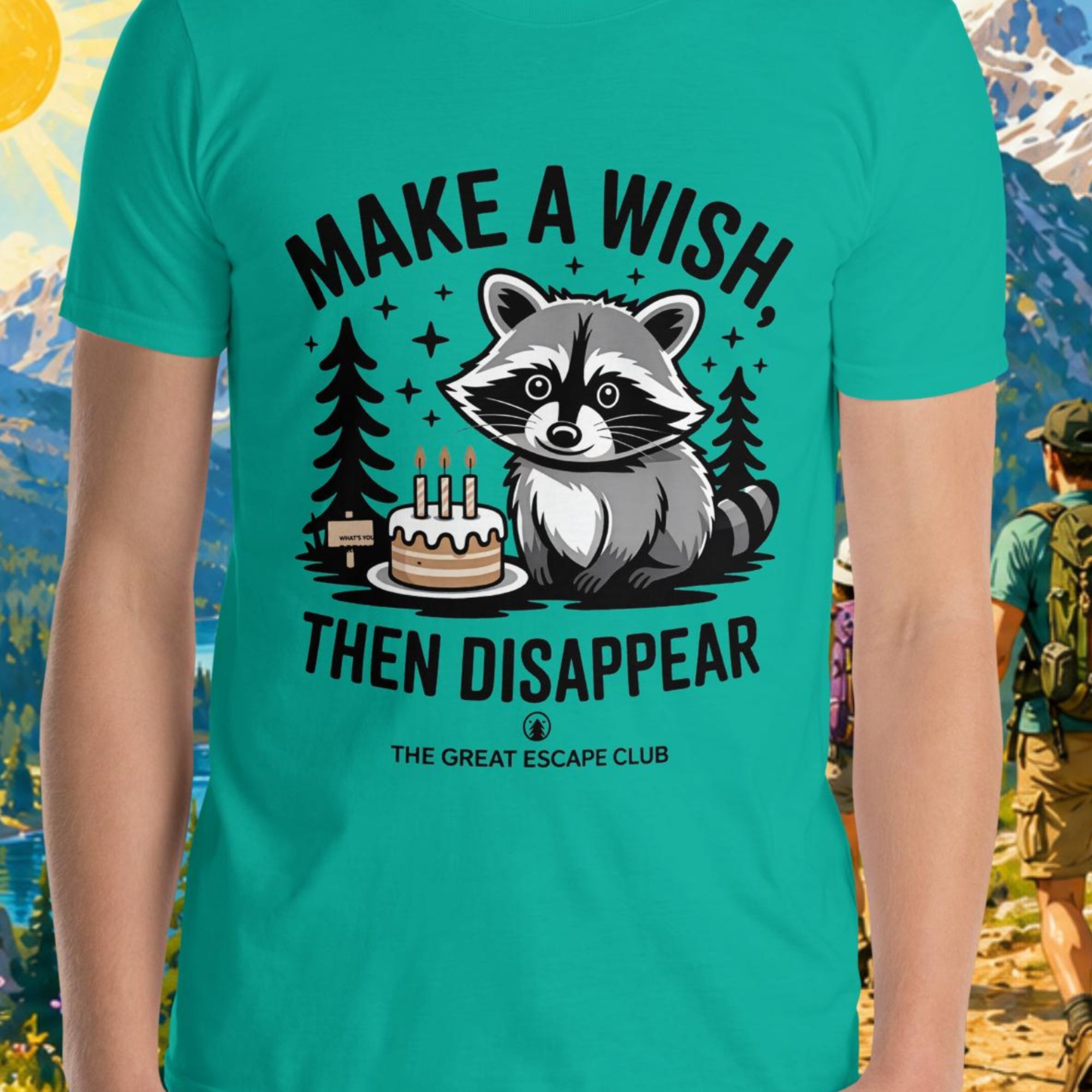 Person wearing a teal t-shirt with a raccoon graphic and text, standing in front of a scenic mountain landscape.